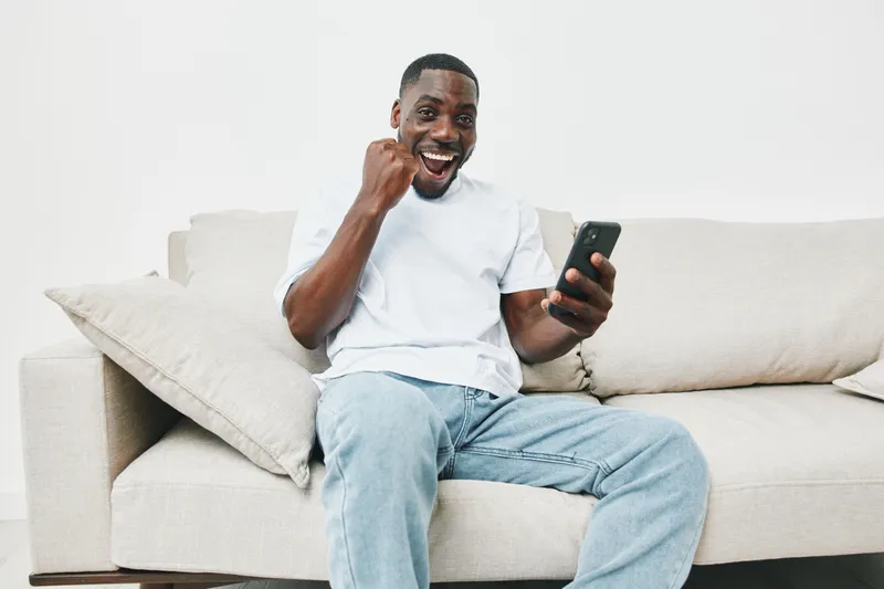 Happy young African American man celebrates success while using smartphone in a modern living room with neutral colors, showcasing joy and excitement