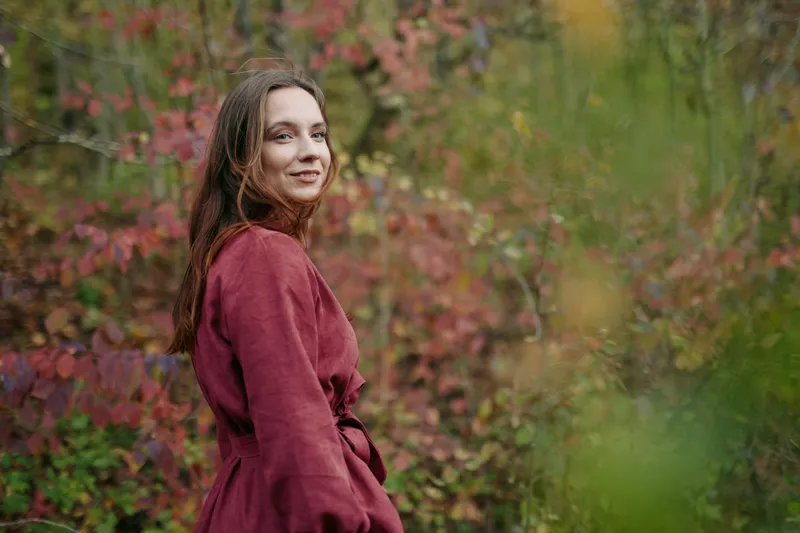 A candid portrait of a young woman in a burgundy coat, standing among autumn foliage, conveying authenticity and sincere mood, quiet confidence, natural pose, subtle smile.