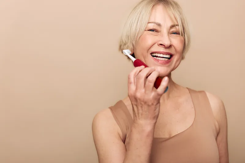 Smiling mature woman with blonde hair holds red electric facial brush near face. Beige background and tank top create warm atmosphere. Concept of skincare, beauty, and self-care for aging skin.