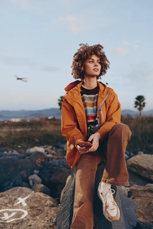 Woman outdoors lifestyle jacket curly sunlight sky rocks handbag fashion portrait, relaxed woman with curly hair in orange jacket and brown pants sits on rocks, warm daylight, distant landscape,