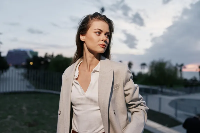 Woman in beige business suit looking away with contemplative expression in outdoor urban environment during sunset with soft natural lighting and light blue sky
