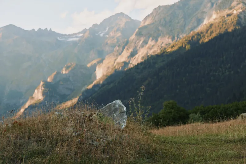 Quiet outdoor landscape with distant mountains, rolling meadow, and rocky hillside, sunlit grasses swaying in a calm natural setting, vibrant yet subtle colors