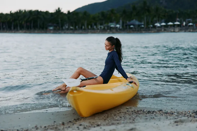 A woman sits on a bright yellow kayak at the edge of a sandy beach, feet in shallow water, gazing toward the calm sea as palm trees line the distant shore.