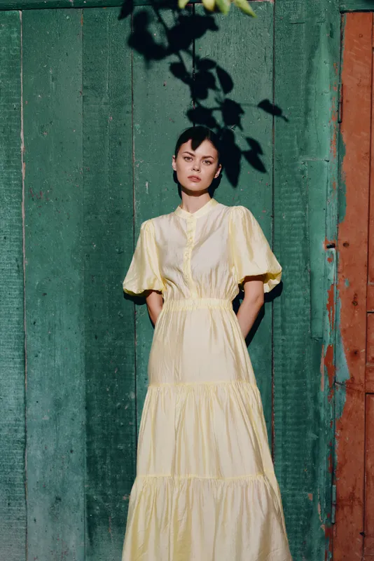 Woman in a pale yellow dress stands by a green wooden wall, with leafy shadows, creating a calm fashion portrait in natural light.