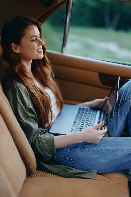 Young woman with long red hair working on a laptop in a car, wearing casual green jacket and jeans, smiling and enjoying the outdoor view.