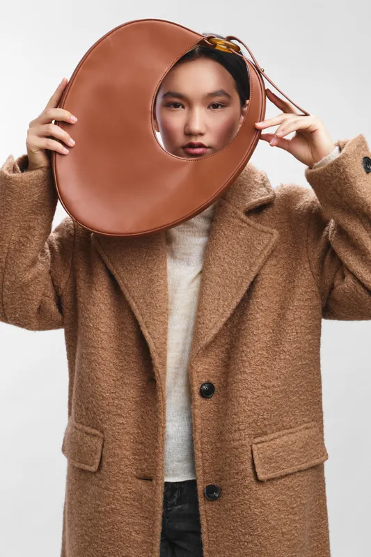 asian woman wearing a warm camel coat holds a large circular leather prop in front of her face, creating a playful fashion portrait with a clean studio backdrop.