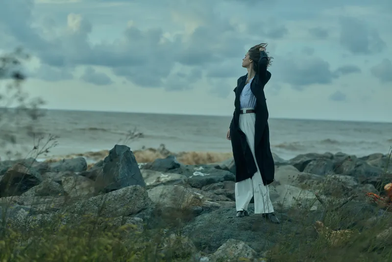 Woman in a long dark coat and white dress stands on a rocky shoreline, gazing at the sea beneath a cloudy sky, wind lifting her hair