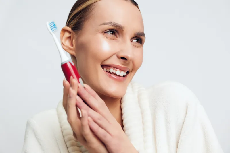 Smiling woman in white bathrobe holding electric toothbrush near face. Close-up on white background. Concept of dental care, hygiene, wellness, and morning routine with natural lighting.