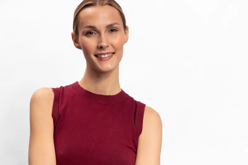 Smiling young woman in sleeveless burgundy top poses against white background. She looks confident and happy with natural makeup and neatly styled hair. Concept of beauty and positivity.
