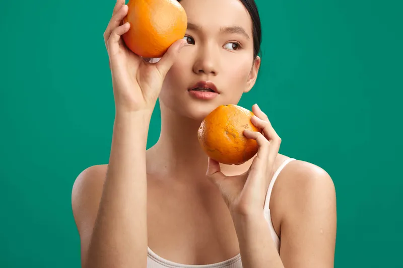 asian woman poses with oranges against a vivid green backdrop, delicate expression, soft lighting highlights smooth skin, casual attire, artistic studio shot emphasizing freshness and playful fruit