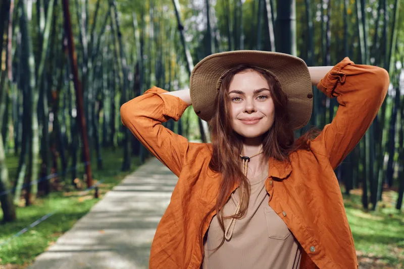 Woman smile hat park pathway portrait outdoor fashion lifestyle image of a young woman wearing a wide brim hat and orange jacket standing on a tree lined path, relaxed and confident.