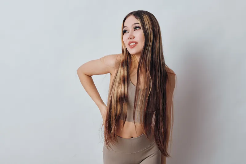Woman with long straight hair and casual sportswear standing with hand on waist looking away isolated on white background. Fitness, style, and beauty concept with confident pose indoors.