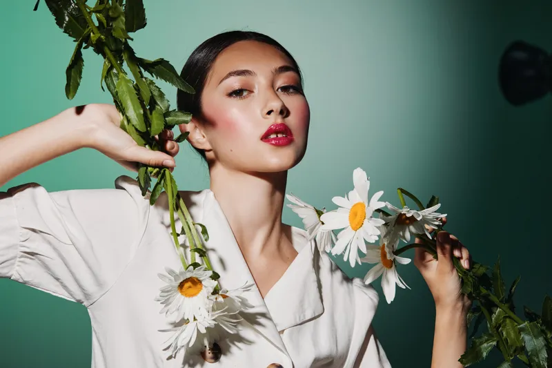 Fashion model with fresh makeup poses holding white daisies wearing a light beige jacket. Studio portrait against green background representing beauty and creativity.