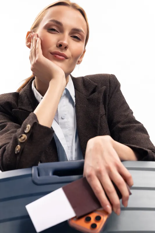 Confident young woman holds suitcase and passport, dressed in stylish business attire. Isolated on white background. Concept for travel, business trip, and modern lifestyle.