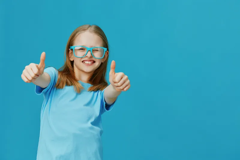 Cheerful girl in blue shirt and glasses giving thumbs up on a bright blue background, showcasing joy and confidence
