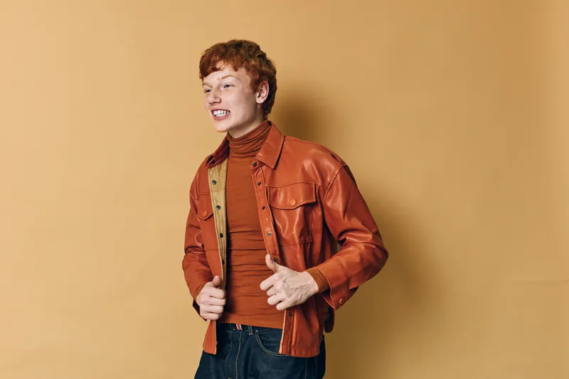 Young man with red hair wearing brown leather jacket and rust turtleneck shirt smiling and adjusting jacket against beige background in studio. Casual style and confident pose.