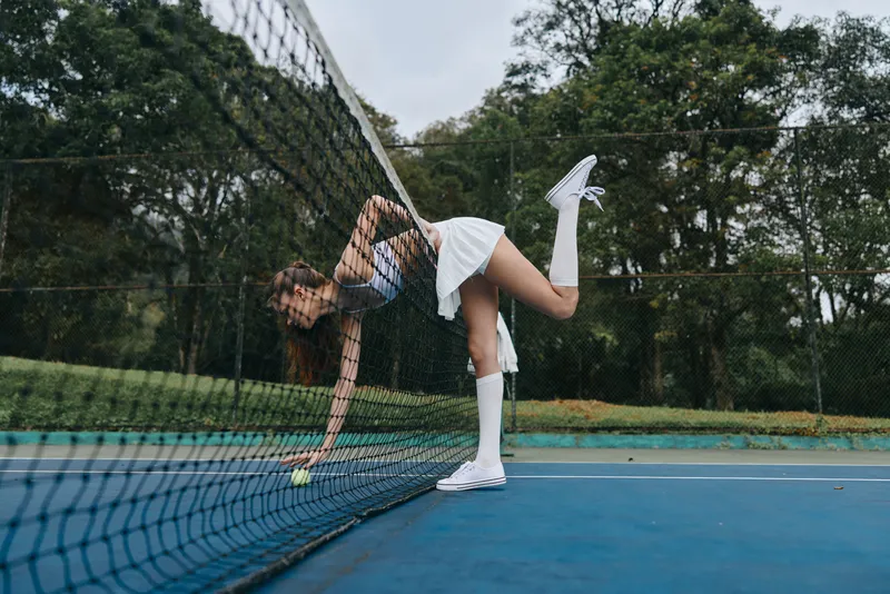 Young athletic woman in white dress playing tennis, showcasing determination, surrounded by lush green trees on a cloudy day on the court