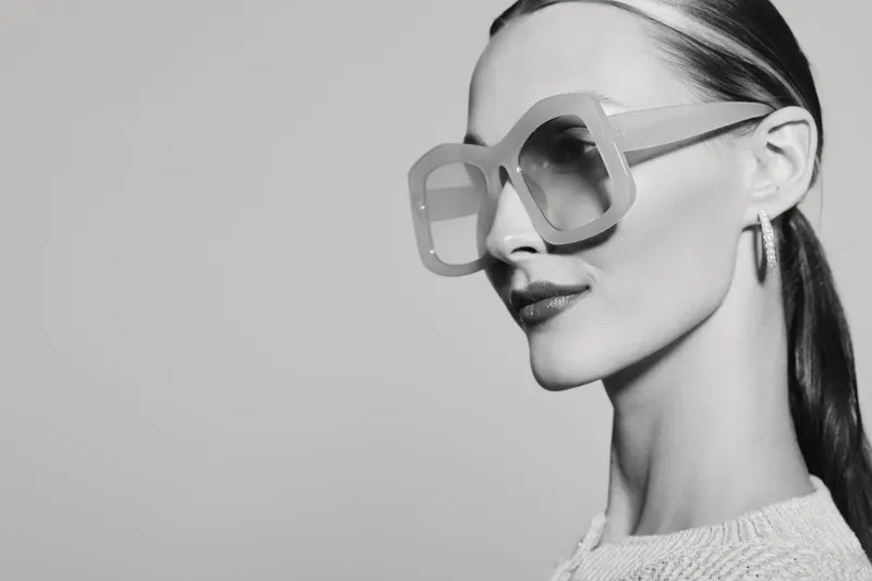 Stylish white woman with pale skin wearing large geometric sunglasses and hoop earrings looks to side, minimalistic monochrome portrait with smooth background and elegant pose.
