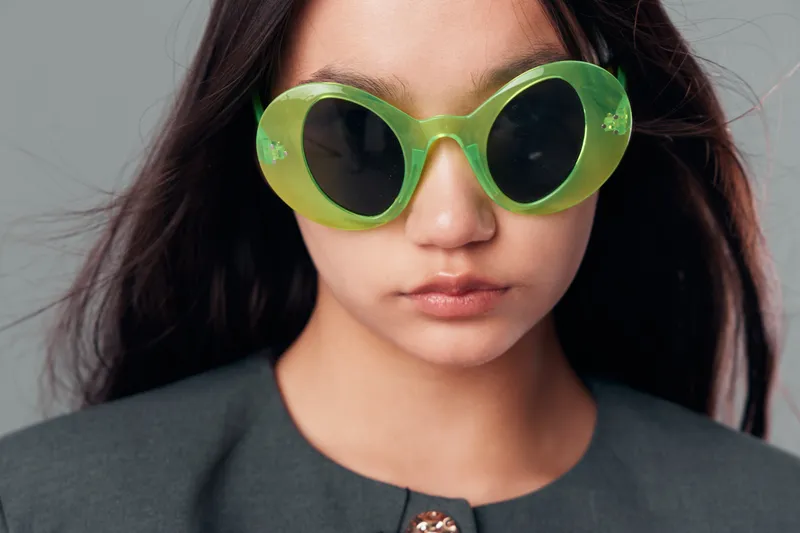 Young woman portrait with green sunglasses looking serious with long dark hair and gray jacket, minimal makeup and neutral background for fashion style and accessories.