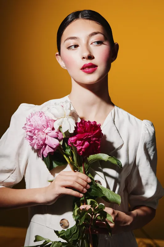 Young woman with elegant makeup and dark sleek hair holds a bouquet of vibrant flowers, wearing a puff sleeve white dress. Studio shot on warm yellow background, beauty fashion concept.