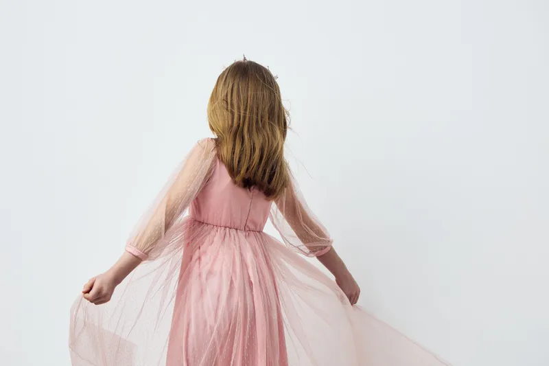 A young girl in a pink dress with flowing tulle, standing gracefully against a minimalist white background, evoking a sense of innocence and elegance