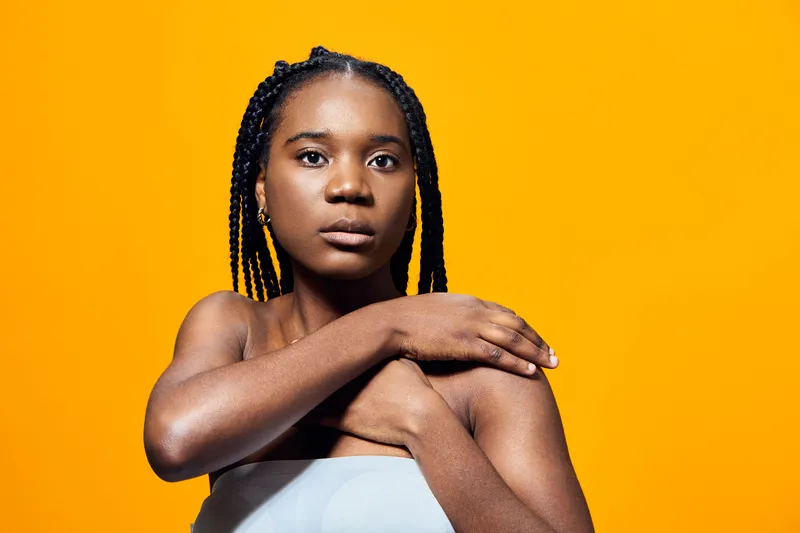Portrait of a young Black woman with braids, wearing a strapless top, expressing confidence against a bright yellow background