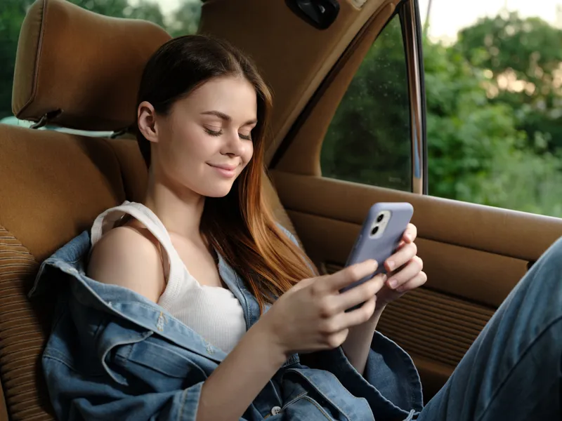 Young woman enjoying her smartphone while relaxing in a car, wearing casual denim, smiling contently in a natural outdoor setting with greenery.