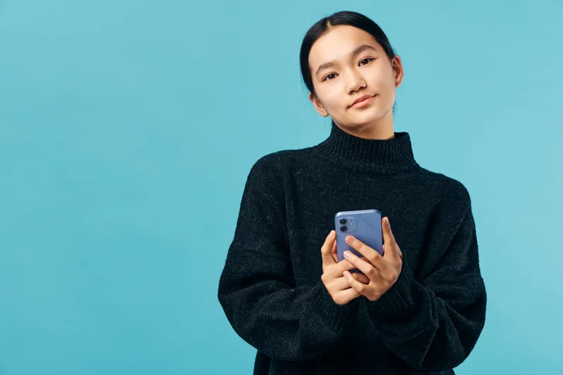Young woman holding smartphone and looking at camera with slight smile in studio. Female with dark hair wearing black sweater poses isolated on blue background for communication or technology.