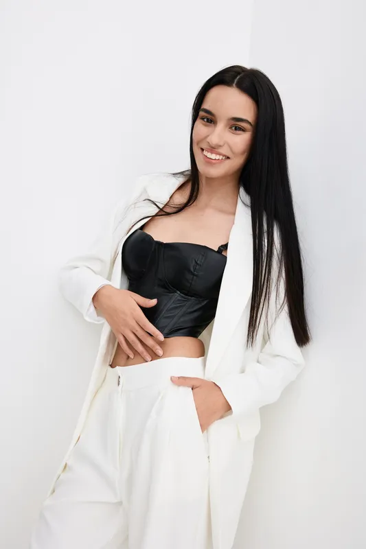 Young smiling woman in white suit and black corset posing confidently with long straight hair against a plain white background, showcasing modern fashion style.