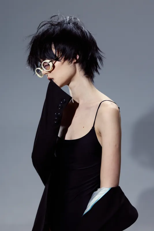 Stylish woman with short hair and glasses wearing black dress and jacket expressing elegance and modern fashion.