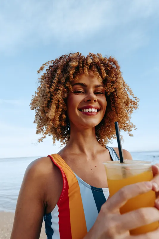 Smiling young woman with curly hair holds a bright orange juice at the beach, wearing a colorful swimsuit as the sea and blue sky create a sunny, refreshing moment for a carefree wellness vibe.