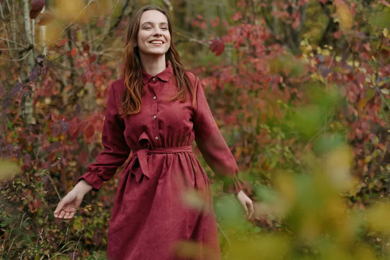 Genuine portrait of a smiling woman in a deep burgundy dress standing among autumn foliage, conveying authentic confidence and natural style in soft outdoor light.