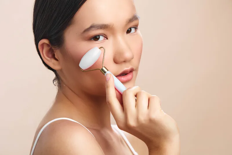 asian woman applies a facial roller in a clean studio, calm and focused, creating a serene skincare moment that conveys wellness, relaxation, beauty, and self care.