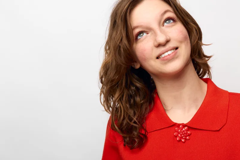 Young woman with curly brown hair wearing red sweater looks up and smiles on white background. Concept of happiness, hope, casual style, and positive emotions.