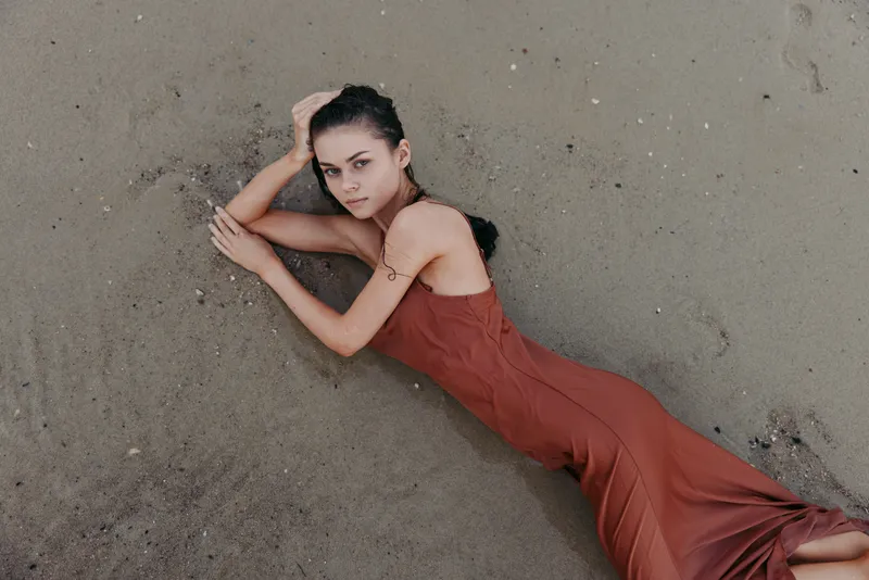 Fashion portrait of a woman lying on sand in a rust dress, with soft light highlighting curves, fabric texture, and the calm shoreline setting.