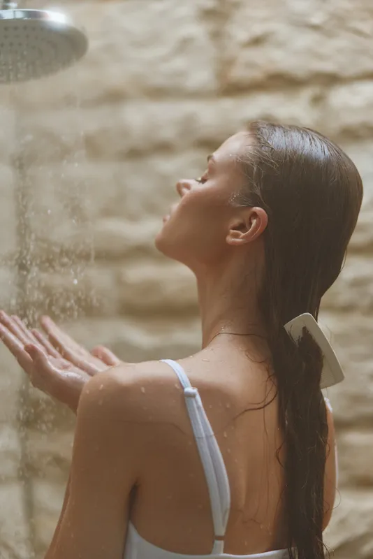 Woman enjoying relaxing shower with water streaming down skin, serene expression, wet hair, neutral stone wall background, peaceful indoor moment.