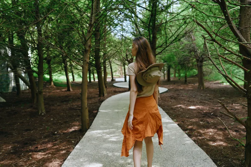 Woman walking on a winding path in a forest, wearing an orange skirt and carrying a hat, summer nature scene with trees and dappled light, peaceful solo outdoor exploration