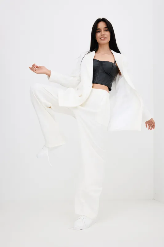 Young woman wearing a white suit and black top poses energetically against a minimalistic white background, showcasing modern fashion and confident style.