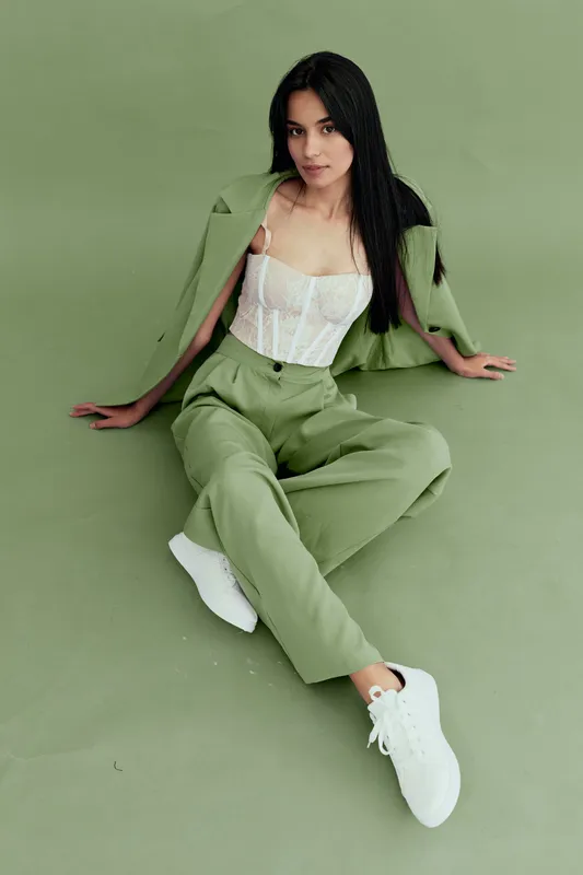 stylish woman wearing green suit and white lace top sits on green floor with white sneakers, posing confidently in minimalistic studio setting