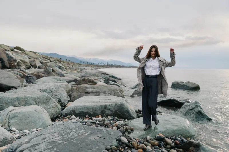Woman in casual outfit walks carefully on large rocks by the sea shore under cloudy sky. The coastal landscape features pebbles, water, and distant mountains creating a calm nature scene.