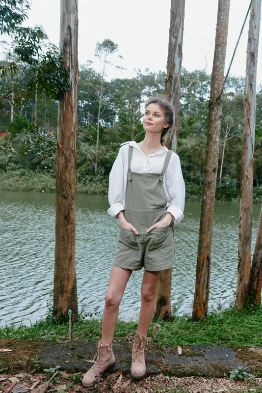 Young woman in olive green overalls stands by a tranquil lake, surrounded by trees, showcasing a relaxed and stylish summer vibe with nature inspired colors