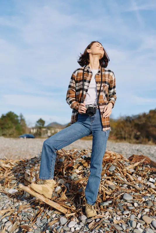 Woman outdoors in casual fashion posing on rocky shoreline, plaid shirt over white tee, denim jeans and boots, looking up at clear blue sky. Natural lifestyle scene suitable for travel, nature and