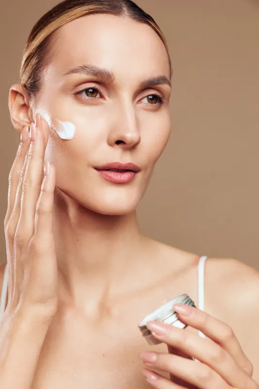 Young woman applies face cream on clean skin holding jar with moisturizer looking away with calm expression isolated on beige background in skincare beauty concept.