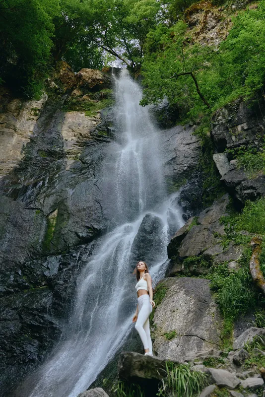 Woman standing by waterfall in lush green forest, wearing white athletic clothes, enjoying nature and fresh air in a peaceful outdoor setting.