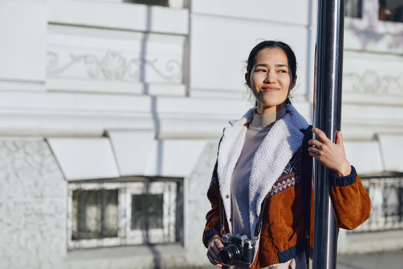 Woman photographer posing beside a pole in an outdoor urban setting, wearing a warm jacket and scarf, holding a camera, daylight street scene, candid and ready for shooting