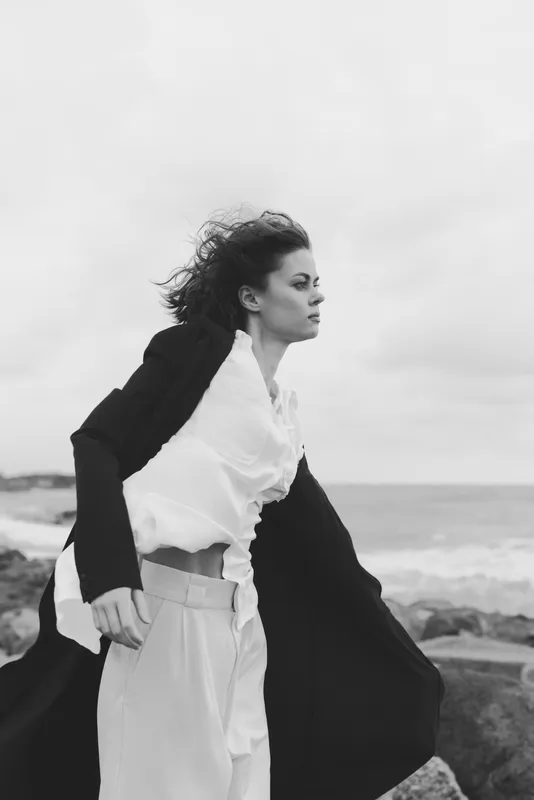 Wind, coat, fashion, seaside, ocean, windblown silhouette of a woman on a windy shoreline in monochrome, dramatic movement and texture
