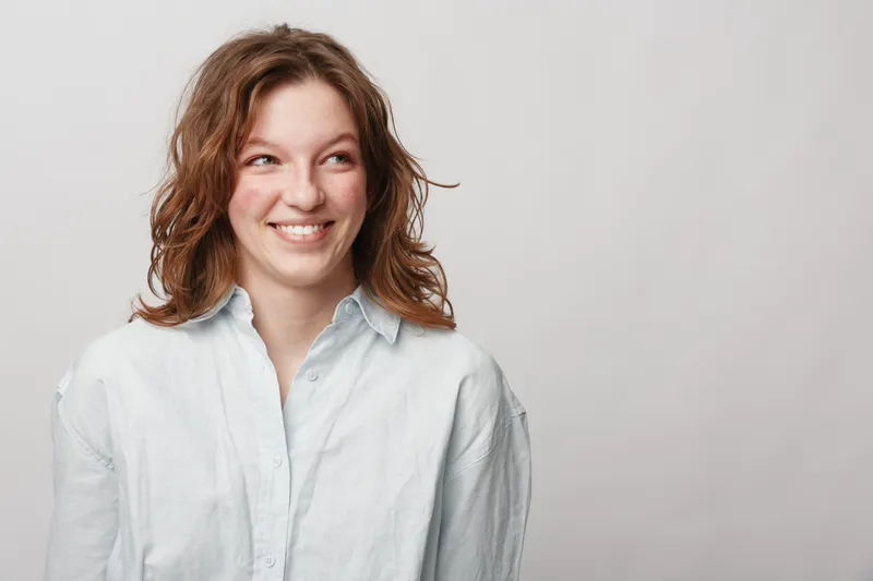 Smiling young woman with wavy brown hair wearing white shirt looks to side with happy expression. Studio portrait on light background. Concept of joy, casual style, and natural beauty.