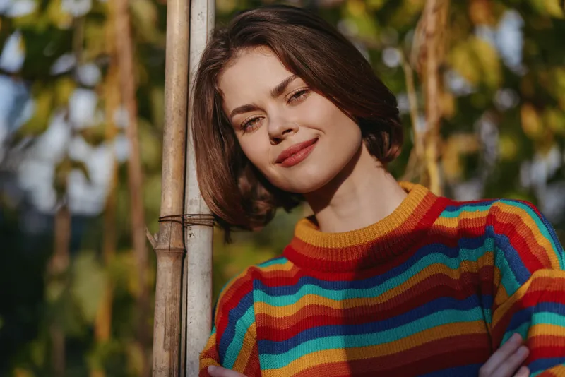 Woman, striped sweater, smiling, leaning, short hair, outdoor: Young woman in her twenties with brown bob smiles gently while leaning on a bamboo pole in a sunlit garden wearing a rainbow striped