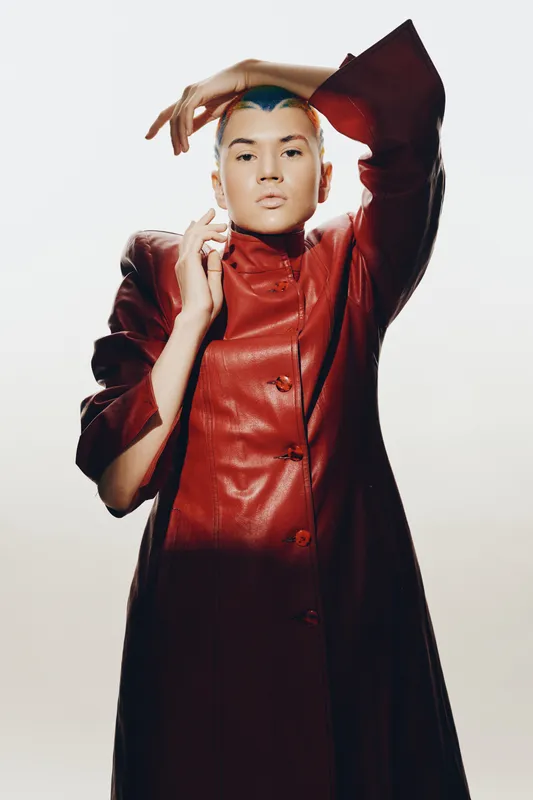 woman with short blue hair wearing a red leather coat posing with raised arm on light background, fashion portrait conveying modern style and confidence
