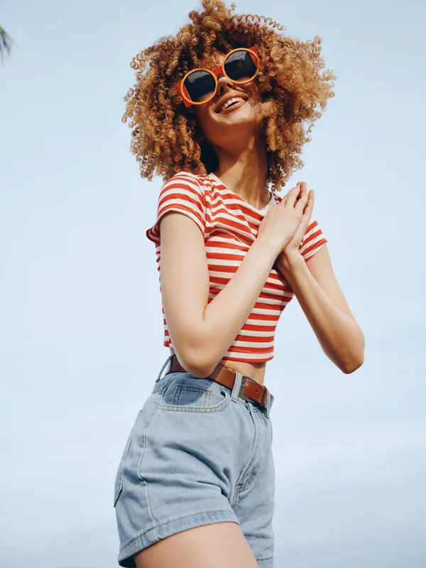 Vibrant portrait of a young woman with curly hair wearing a red striped crop top and denim shorts, posing confidently against a clear blue sky, and a joyful expression.